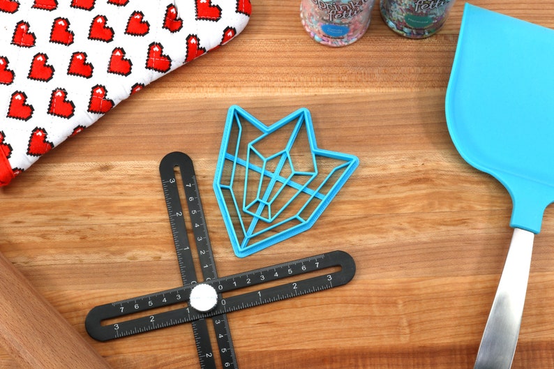 May include: A blue plastic geometric cookie cutter in the shape of an arrow on a wooden surface with a black ruler and a blue spatula.