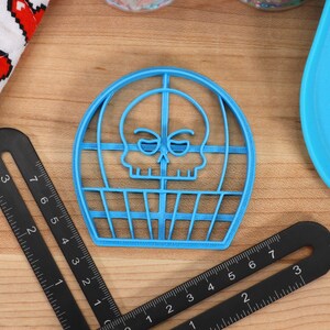 May include: A blue plastic cookie cutter shaped like a skull in a cage. The skull has a cartoonish expression with raised eyebrows and a small smile.
