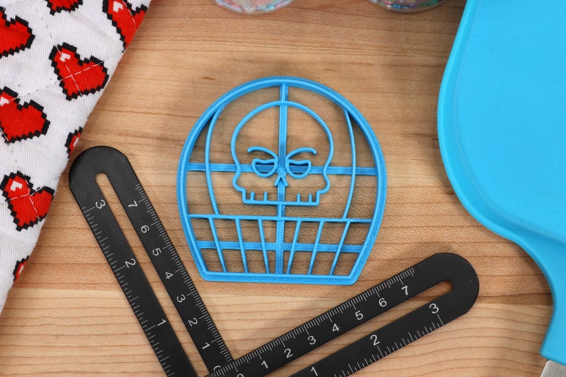 May include: A blue plastic cookie cutter shaped like a skull in a cage. The skull has a cartoonish expression with raised eyebrows and a small smile.