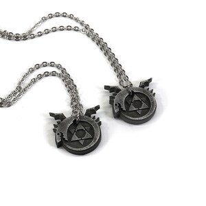 K&ouml;nnte beinhalten: Zwei silberne Halsketten mit passenden Anh&auml;ngern, die ein kreisf&ouml;rmiges Design in Schwarz und Silber mit einem Dreieck darin zeigen. Das Design erinnert an ein Symbol aus dem Anime Fullmetal Alchemist.