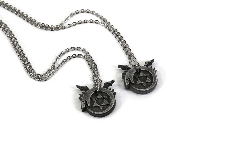K&ouml;nnte beinhalten: Zwei silberne Halsketten mit passenden Anh&auml;ngern, die ein kreisf&ouml;rmiges Design in Schwarz und Silber mit einem Dreieck darin zeigen. Das Design erinnert an ein Symbol aus dem Anime Fullmetal Alchemist.