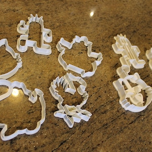 May include: A collection of white plastic cookie cutters in various shapes, including a cactus, a cat, and other cartoon-like figures. The cutters are arranged on a speckled brown countertop.