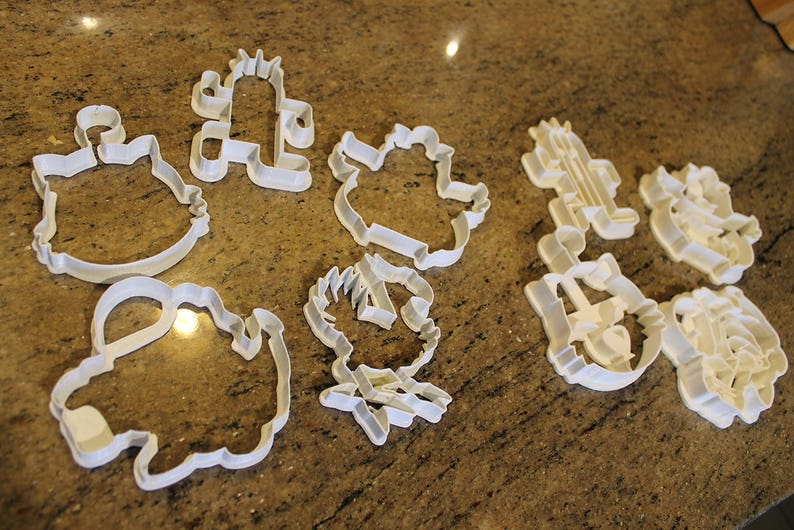 May include: A collection of white plastic cookie cutters in various shapes, including a cactus, a cat, and other cartoon-like figures. The cutters are arranged on a speckled brown countertop.