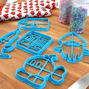 May include: A set of blue plastic cookie cutters shaped like a banana, a square, a squid, a character with a large head, and a character with a large head and a hat. The cutters are on a wooden surface with a rolling pin and a container of sprinkles.