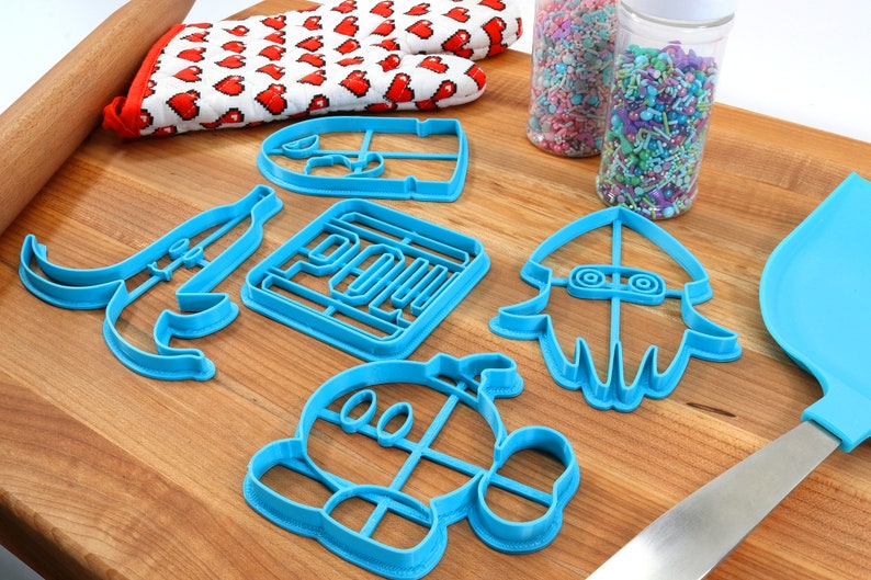 May include: A set of blue plastic cookie cutters shaped like a banana, a square, a squid, a character with a large head, and a character with a large head and a hat. The cutters are on a wooden surface with a rolling pin and a container of sprinkles.