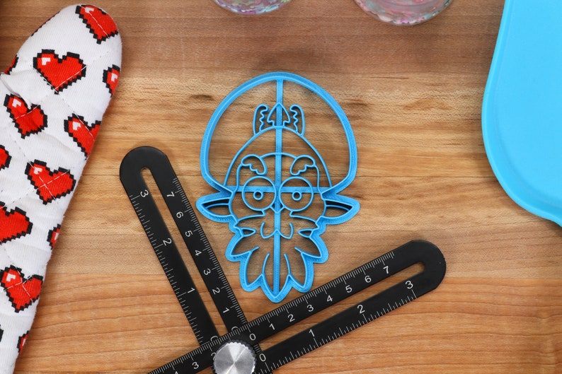 May include: Blue plastic cookie cutter shaped like a cartoon character with a beard and a hat. The character has large eyes and a wide smile.