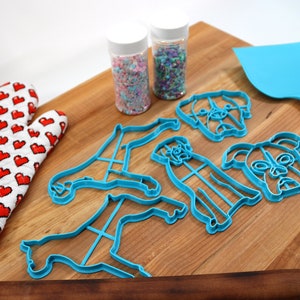 May include: A set of blue plastic dog-shaped cookie cutters on a wooden cutting board. There are two jars of sprinkles, a red and white oven mitt, and a blue spatula.