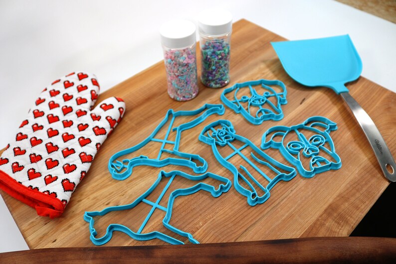 May include: A set of blue plastic dog-shaped cookie cutters on a wooden cutting board. There are two jars of sprinkles, a red and white oven mitt, and a blue spatula.
