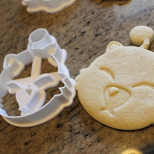 May include: A close-up of a bear-shaped cookie next to a white plastic cookie cutter. The cookie is light brown and has a smiling face. The cutter is in the shape of the bear's face.