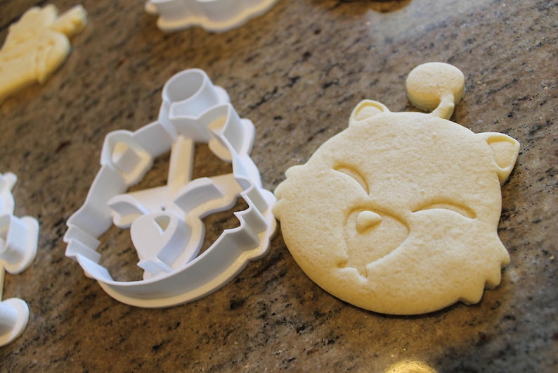 May include: A close-up of a bear-shaped cookie next to a white plastic cookie cutter. The cookie is light brown and has a smiling face. The cutter is in the shape of the bear's face.