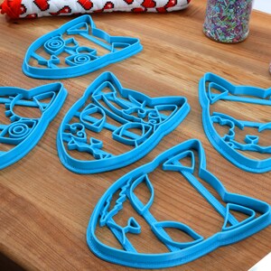 K&ouml;nnte beinhalten: F&uuml;nf blaue, plastik Katzen-f&ouml;rmige Ausstecher auf einer Holzoberfl&auml;che. Die Ausstecher haben detaillierte Merkmale, einschlie&szlig;lich Augen, Schnurrhaare und Streifen.