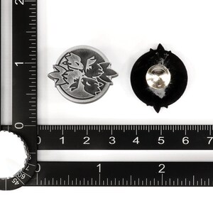 Op de afbeelding: Twee metalen pinnen, een met een zilveren afwerking en een bloemmotief, de andere met een zwarte afwerking en een stervormig motief. Beide pinnen zijn ongeveer 2,5 cm in diameter.