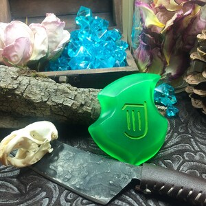 May include: A green, translucent, shield-shaped pendant with a stylized harp design. The pendant is lying on a dark, patterned surface with a skull, a knife, and dried flowers.