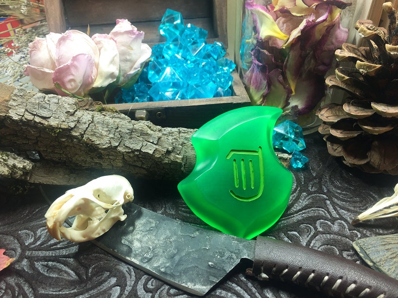 May include: A green, translucent, shield-shaped pendant with a stylized harp design. The pendant is lying on a dark, patterned surface with a skull, a knife, and dried flowers.