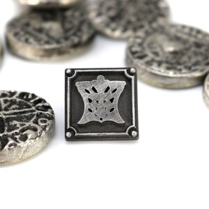 May include: A silver metal square pin with a detailed design of a crown inside. The pin is surrounded by several silver coins with intricate designs.