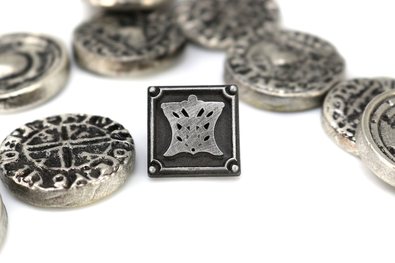 May include: A silver metal square pin with a detailed design of a crown inside. The pin is surrounded by several silver coins with intricate designs.