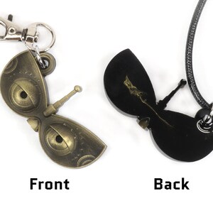 May include: A gold-colored metal butterfly pendant with a detailed design and a black cord necklace. The pendant has a small, silver-colored hook on the back. The front of the pendant has a detailed design with two large eyes. The back of the pendant is black and has a smaller, silver-colored hook.