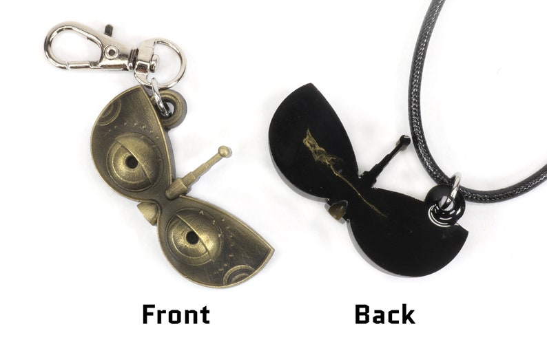 May include: A gold-colored metal butterfly pendant with a detailed design and a black cord necklace. The pendant has a small, silver-colored hook on the back. The front of the pendant has a detailed design with two large eyes. The back of the pendant is black and has a smaller, silver-colored hook.
