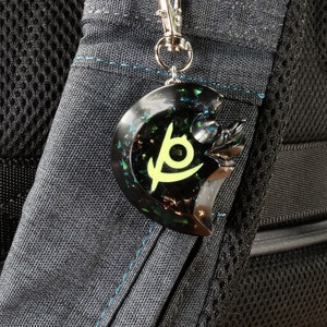 May include: A black and green resin keychain with a crescent moon shape and a stylized symbol in the center. The symbol is a yellow crescent moon with a green eye inside.