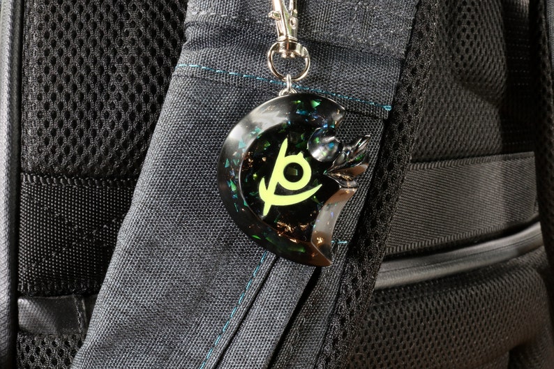 May include: A black and green resin keychain with a crescent moon shape and a stylized symbol in the center. The symbol is a yellow crescent moon with a green eye inside.