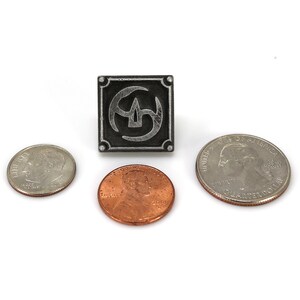 May include: A silver metal pin with a black and silver design. The pin is square shaped and features a stylized symbol with a circle and three curved lines. The pin is about the size of a US quarter.