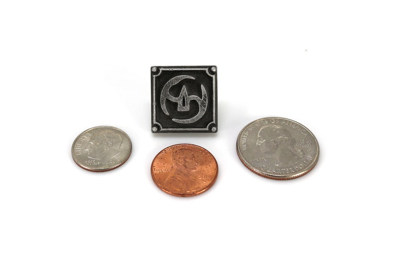 May include: A silver metal pin with a black and silver design. The pin is square shaped and features a stylized symbol with a circle and three curved lines. The pin is about the size of a US quarter.