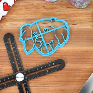 May include: A blue plastic cookie cutter shaped like a dog's head. The cutter is on a wooden surface with a black ruler and a blue spatula.