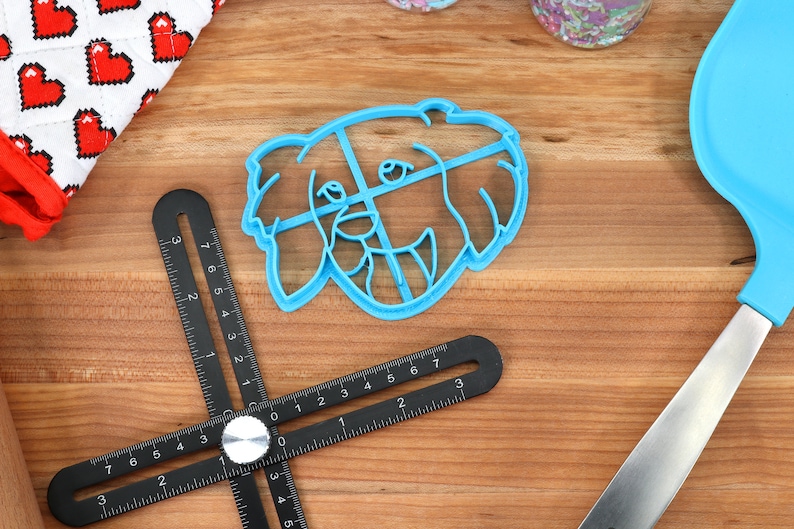 May include: A blue plastic cookie cutter shaped like a dog's head. The cutter is on a wooden surface with a black ruler and a blue spatula.
