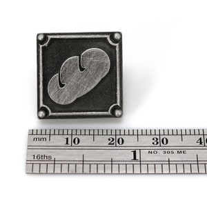 May include: A silver-colored metal pin with a black background. The pin features a stylized image of a loaf of bread. The pin is approximately 2 cm wide.