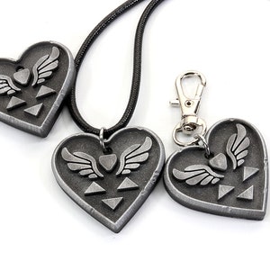 Puede incluir: Tres colgantes de coraz&oacute;n plateados con un s&iacute;mbolo alado y un dise&ntilde;o triangular. Los colgantes est&aacute;n en un collar de cord&oacute;n negro y un llavero plateado.