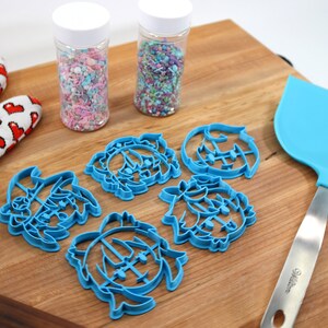 K&ouml;nnte beinhalten: Ein Schneidebrett aus Holz mit f&uuml;nf blauen Plastik-Ausstechern in Form von Comic-Gesichtern, zwei Gl&auml;sern mit bunten Streuseln, einem blauen Silikonspatel und einem roten und wei&szlig;en Ofenhandschuh.