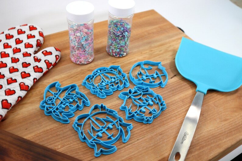 K&ouml;nnte beinhalten: Ein Schneidebrett aus Holz mit f&uuml;nf blauen Plastik-Ausstechern in Form von Comic-Gesichtern, zwei Gl&auml;sern mit bunten Streuseln, einem blauen Silikonspatel und einem roten und wei&szlig;en Ofenhandschuh.