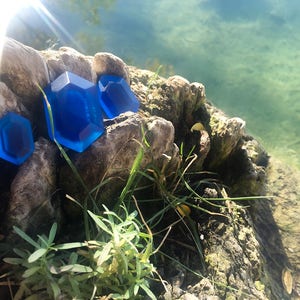 May include: Three blue gemstone-shaped objects on a rock near a body of water. The gemstones are different sizes and shapes.