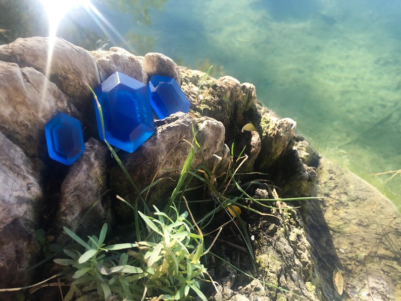 May include: Three blue gemstone-shaped objects on a rock near a body of water. The gemstones are different sizes and shapes.