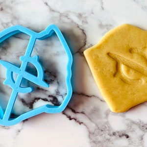 May include: A blue plastic cookie cutter shaped like a compass with a crosshair design. A cookie is shown next to the cutter, with the compass design imprinted on it.