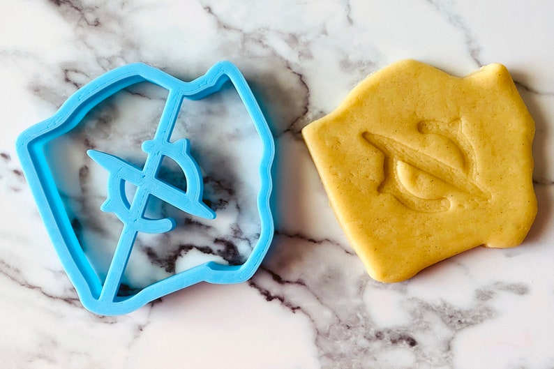 May include: A blue plastic cookie cutter shaped like a compass with a crosshair design. A cookie is shown next to the cutter, with the compass design imprinted on it.