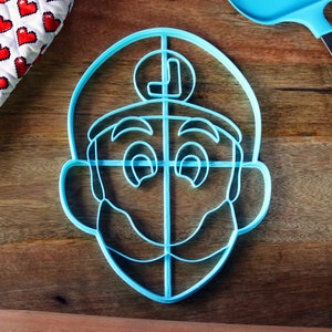 May include: A light blue plastic cookie cutter in the shape of a cartoon character's face, with a mustache and a letter "L" on the hat. A blue spatula and a heart-patterned oven mitt are also visible.