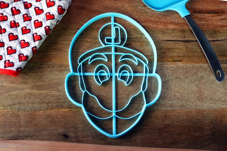 May include: A light blue plastic cookie cutter in the shape of a cartoon character's face, with a mustache and a letter "L" on the hat. A blue spatula and a heart-patterned oven mitt are also visible.