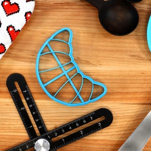 May include: A light blue croissant-shaped cookie cutter, a black adjustable angle ruler, a white oven mitt with red pixelated hearts, black measuring spoons, and a blue spatula are arranged on a wooden surface.