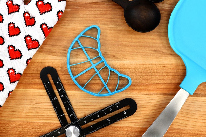May include: A light blue croissant-shaped cookie cutter, a black adjustable angle ruler, a white oven mitt with red pixelated hearts, black measuring spoons, and a blue spatula are arranged on a wooden surface.