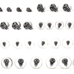 May include: A collection of black and gray ear gauges and plugs. The gauges feature a stylized skull design with three eye holes and a claw-like structure. The plugs are clear with a skull graphic. The items are arranged in rows on a white background.
