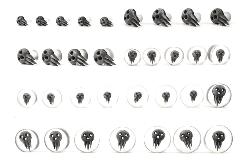 May include: A collection of black and gray ear gauges and plugs. The gauges feature a stylized skull design with three eye holes and a claw-like structure. The plugs are clear with a skull graphic. The items are arranged in rows on a white background.