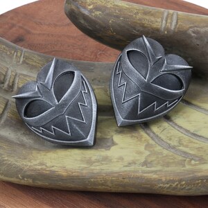 May include: Two silver heart-shaped pendants with a menacing, toothy grin design. The pendants are on a wooden surface.