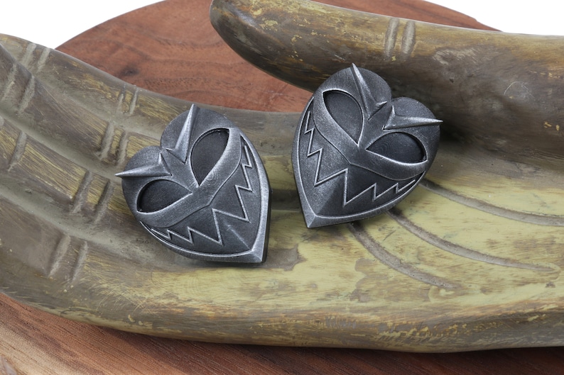 May include: Two silver heart-shaped pendants with a menacing, toothy grin design. The pendants are on a wooden surface.