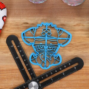 May include: Blue plastic cookie cutter shaped like a cartoon character wearing a hat and glasses. The character has a large mustache and a flower on their hat.