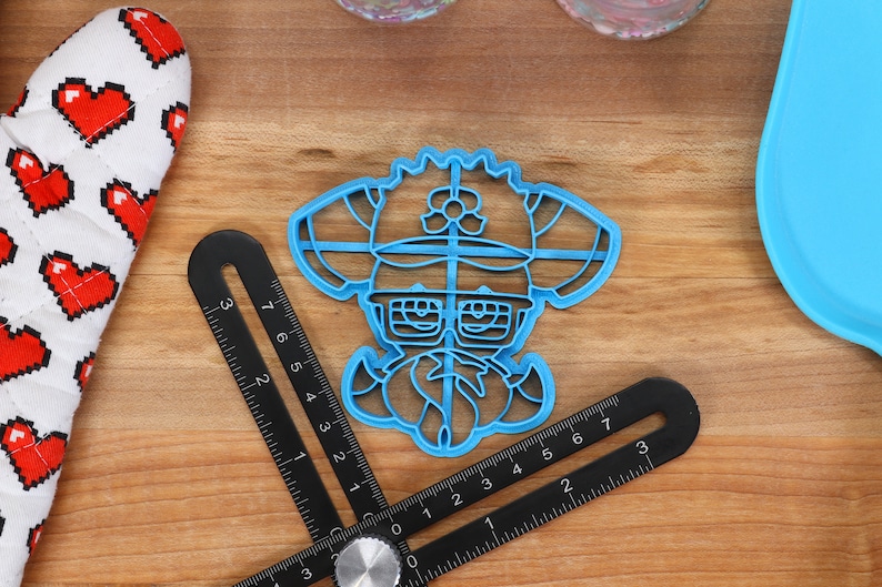 May include: Blue plastic cookie cutter shaped like a cartoon character wearing a hat and glasses. The character has a large mustache and a flower on their hat.