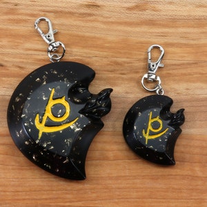 May include: Two black keychains with gold flecks and a yellow symbol. The larger keychain is on the left and the smaller keychain is on the right.