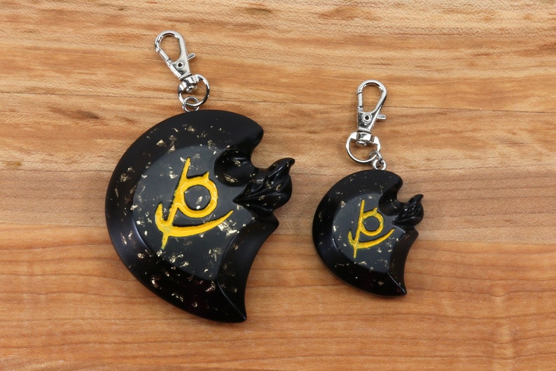 May include: Two black keychains with gold flecks and a yellow symbol. The larger keychain is on the left and the smaller keychain is on the right.