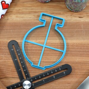 May include: A blue plastic cookie cutter shaped like a circle with a cross inside. The cutter is on a wooden surface.