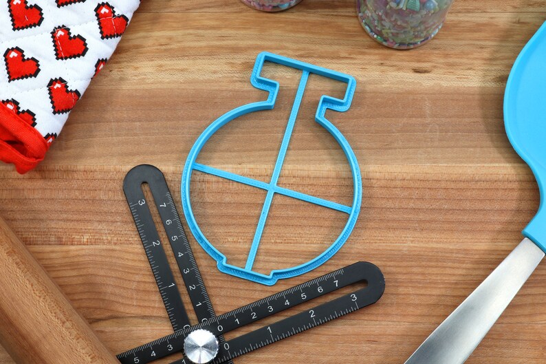 May include: A blue plastic cookie cutter shaped like a circle with a cross inside. The cutter is on a wooden surface.